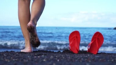 Dişi bacaklar yavaşça siyah kumun üzerinden okyanusa gider. Ön planda kırmızı parmak arası terlikler. Ağır çekim. Kanarya Sahili, Tenerife. Mayıs 2019.