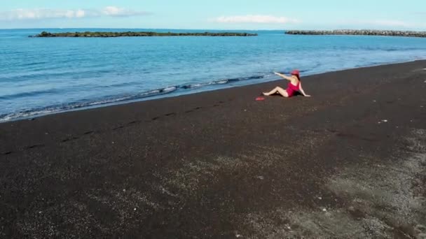 Een Vrouw Een Rood Badpak Hoed Zit Een Zwart Zandstrand — Stockvideo