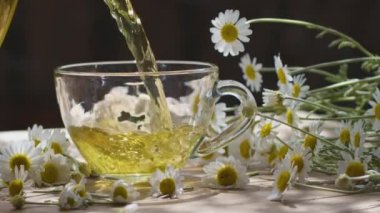 Papatya çayı şeffaf bir çaydanlıktan cam bir fincana dökülür. Eylem, yaz aylarında kırsal bir terasta güneş ışınları eşliğinde gerçekleşiyor. Yakınlarda bir ilaç papatyasının çiçekleri var..