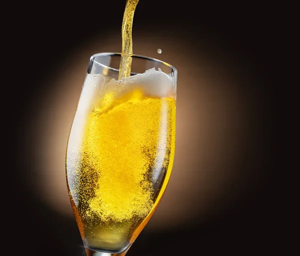 Jet of beer is poured into a beer glass, causing a lot of bubbles and foam. Dark brown background.