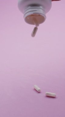 Beyaz kapsüller ya da plastik bir şişeden pembe arka plana dökülen haplar. İlaç, sağlık, takviye ve ilaç tedavisi kavramı.