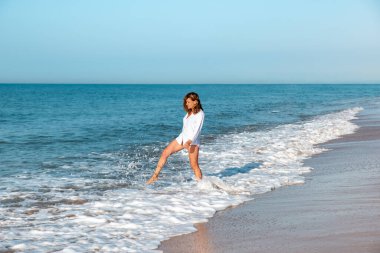 Beyaz gömlekli bir kadın kumsalda dalgadan atlıyor.