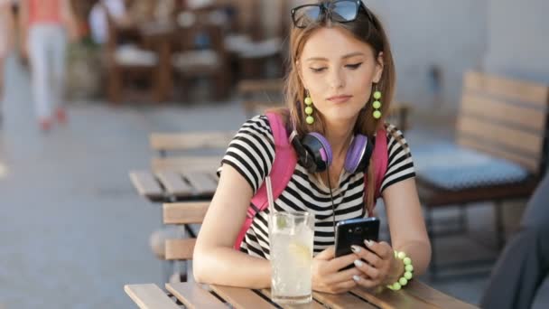 美丽的白种女孩使用智能手机在户外咖啡馆 想什么文本讲口香糖喝冷饮 穿着华丽的黄色首饰和粉红色的背包在温暖的夏日午后 — 图库视频影像