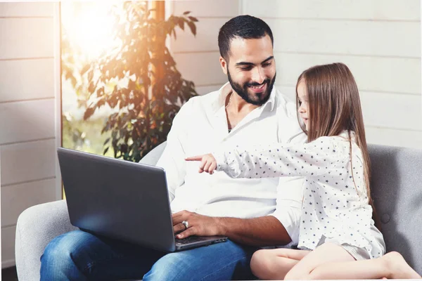 Bearded multicultural father working with laptop and laughing to his little cute daughter in the comfortable living-room
