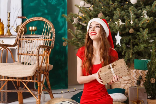Hilarante navidad mujer usando santa sombrero sentado y sosteniendo un regalo —  Fotos de Stock
