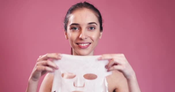 Chica Sonriente Sosteniendo Máscara Facial Mirando Través Máscara Fondo Rosa — Vídeo de stock