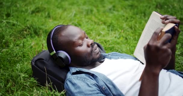 Афроамериканец, лежащий на лужайке с книгой — стоковое видео