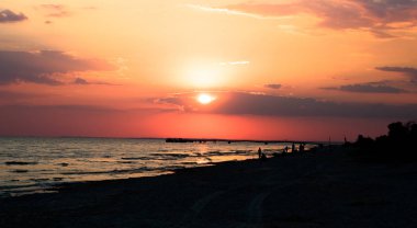 Sahilde kırmızı günbatımı. İnsanlar siluetleri ve martı sahilde. İskelede manzarası