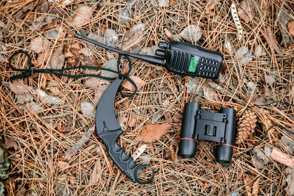 binoculars, radio set and karambit knife on the ground covered with pine needles