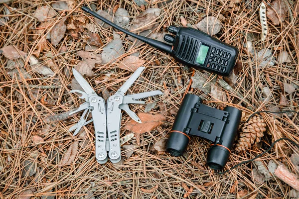 binoculars, radio set and multitool knife on the ground covered with pine needles