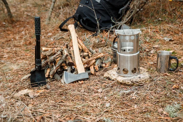 Campingholzofen und -utensilien, Axt und Sägeschaufel in der Nähe eines Feuerwaldes, im Hintergrund ein Rucksack — Stockfoto