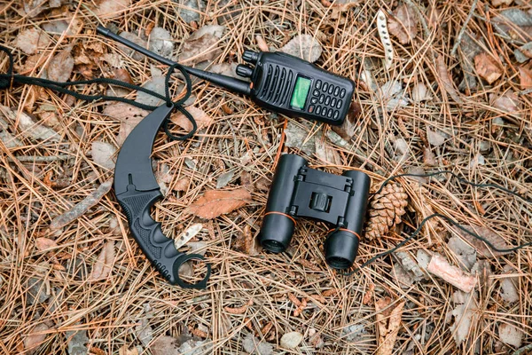 binoculars, radio set and karambit knife on the ground covered with pine needles