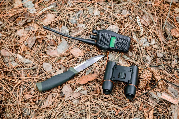 binoculars, radio set and hunting knife on the ground covered with pine needles