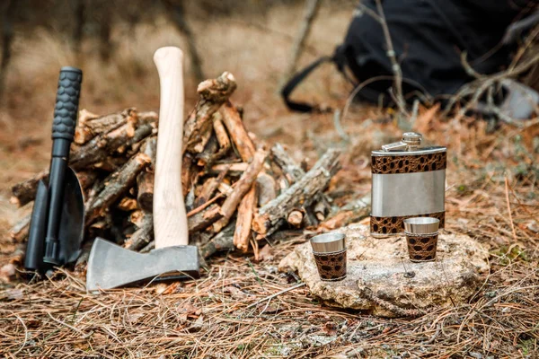 flask with glasses, camping woodstove and utensils, axe and sapper shovel near a firewoods, backpack on the background