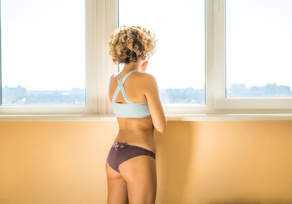Beautiful caucasian curly female in underwear in front of window. orange light background — Stock Photo, Image