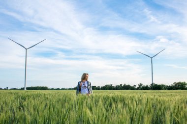 Genç ve çekici bir kadın buğday tarlasında rüzgâr türbini jeneratörlerinin yanında, gün batımında.