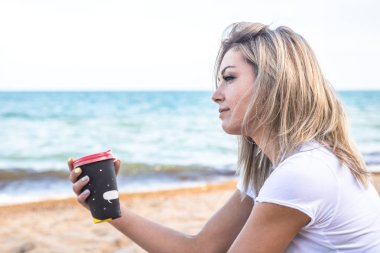 Genç, çekici, sarışın, kahve fincanı olan kadın denize bakıyor.