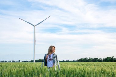 Genç ve çekici bir kadın buğday tarlasında rüzgâr türbini jeneratörlerinin yanında, gün batımında.