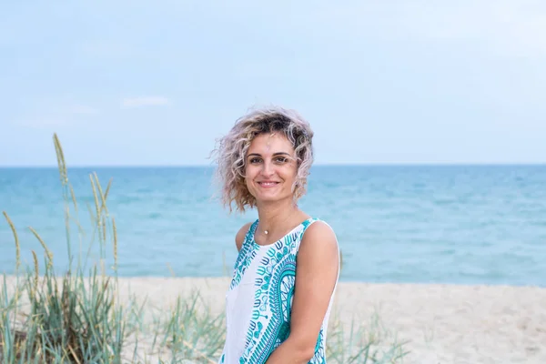 Vacker ung kvinna med lockigt hår i vit klänning med trasery är glad att vara på stranden. Hav och gräs på bakgrunden — Stockfoto