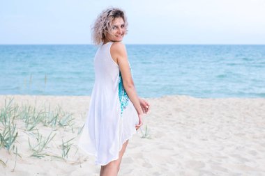 trasery ile beyaz elbise kıvırcık saçlar ile güzel genç kadın plajda olmaktan mutluluk duyacaktır. Arka planda okyanus ve çim