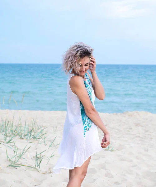 Beautiful young woman with curly hairs in white dress with trasery is happy to be on the beach. Ocean and grass on the background — Stock Photo, Image
