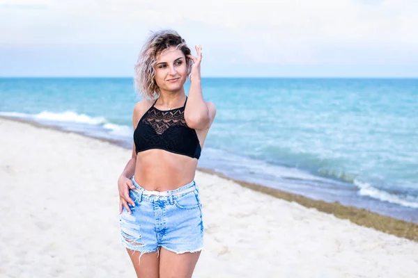 Beautiful young woman with curly hairs in denim shorts and swimsuit is happy to be on the beach near the ocean. Ocean and grass on the background — Stock Photo, Image