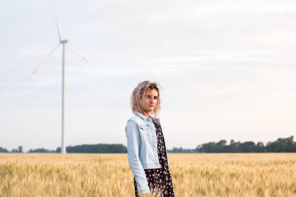 young tanned curly haired woman in denim jacket and dress on the field of ripened wheat, sunset time