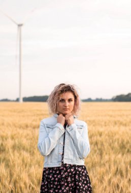 Kotlu ceketli, buğday tarlasında giyinmiş, günbatımında bronzlaşmış kıvırcık saçlı genç bir kadın.