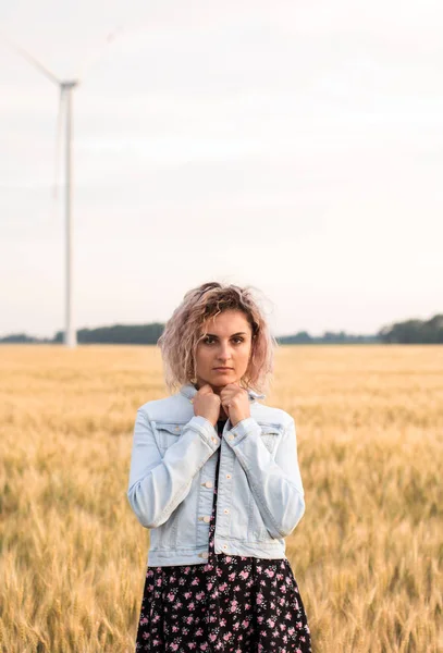 Kotlu ceketli, buğday tarlasında giyinmiş, günbatımında bronzlaşmış kıvırcık saçlı genç bir kadın.