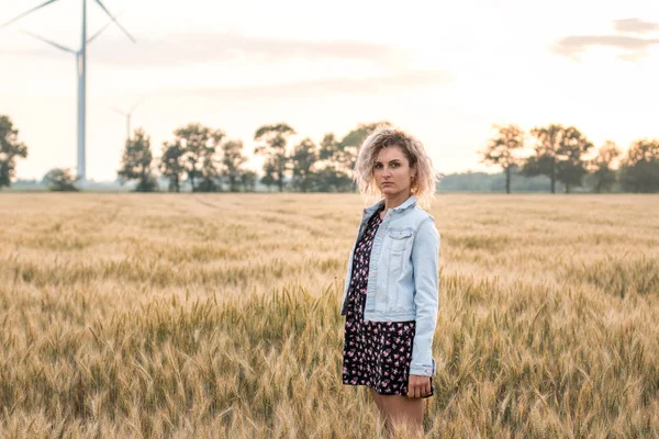 Kotlu ceketli, buğday tarlasında giyinmiş, günbatımında bronzlaşmış kıvırcık saçlı genç bir kadın.