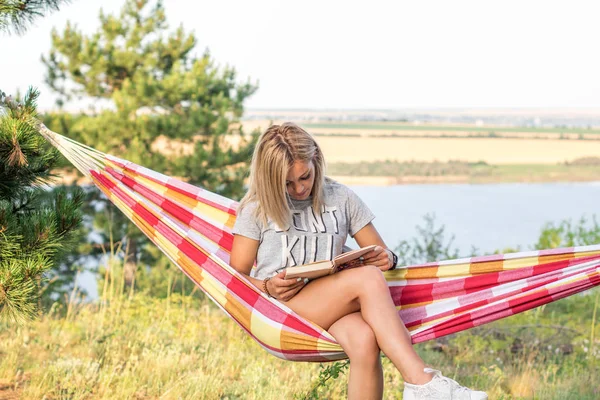 Młoda atrakcyjna biała kobieta czyta książkę w hamaku w lesie, jezioro na tle — Zdjęcie stockowe