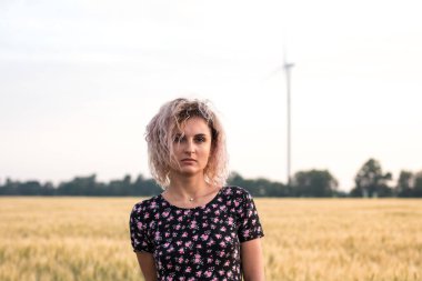 elbiseli genç tabaklanmış kıvırcık saçlı kadın olgunlaşmış buğday alanında olmaktan mutlu, arka planda rüzgar türbini