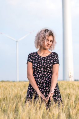 elbiseli genç tabaklanmış kıvırcık saçlı kadın olgunlaşmış buğday alanında olmaktan mutlu, arka planda rüzgar türbini