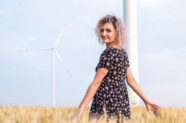 elbiseli genç tabaklanmış kıvırcık saçlı kadın olgunlaşmış buğday alanında olmaktan mutlu, arka planda rüzgar türbini