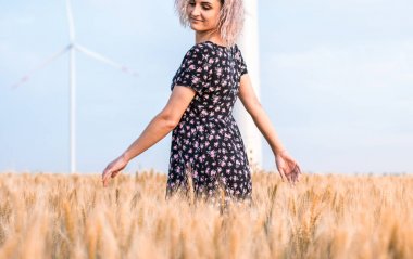 elbiseli genç tabaklanmış kıvırcık saçlı kadın olgunlaşmış buğday alanında olmaktan mutlu, arka planda rüzgar türbini