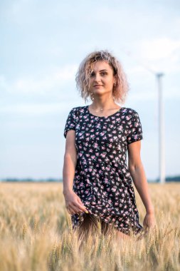 elbiseli genç tabaklanmış kıvırcık saçlı kadın olgunlaşmış buğday alanında olmaktan mutlu, arka planda rüzgar türbini