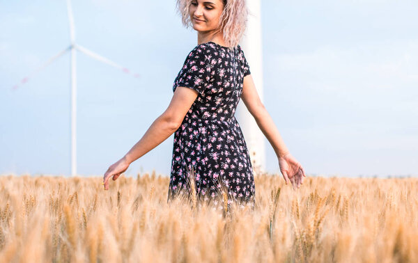 young tanned curly haired woman in dress is happy to be on the field of ripened wheat, wind turbine on the background