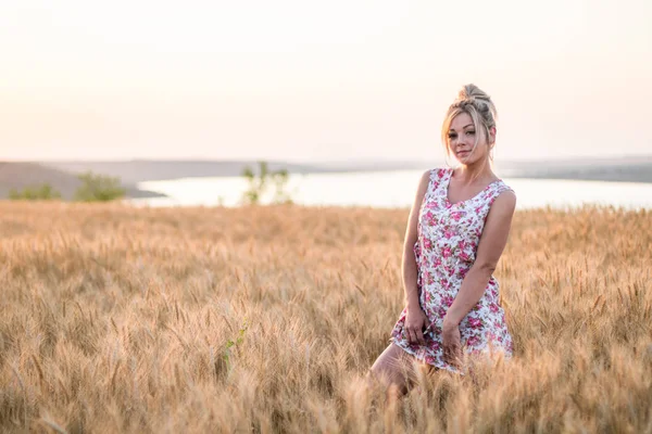 Jeune femme blonde rurale séduisante en robe sur le champ de blé jaune au coucher du soleil, lac en arrière-plan — Photo