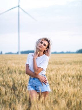 buğday alanında beyaz t-kısa ve denim şort genç tabaklanmış kıvırcık saçlı kadın, arka planda rüzgar türbini