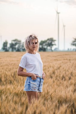 buğday alanında beyaz t-kısa ve denim şort genç tabaklanmış kıvırcık saçlı kadın, arka planda rüzgar türbini