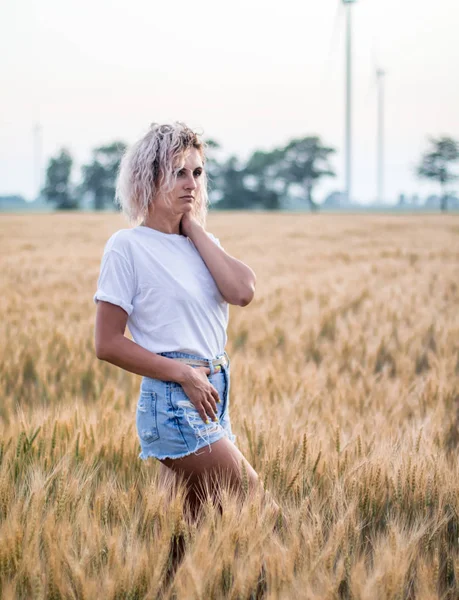 Mujer joven bronceada de pelo rizado en pantalones cortos blancos y pantalones cortos de mezclilla en el campo de trigo, turbina de viento en el fondo —  Fotos de Stock