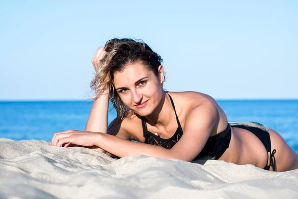 Gebräunte junge attraktive Frau mit lockigem Haar im schwarzen Badeanzug am Strand liegend im Sand — Stockfoto