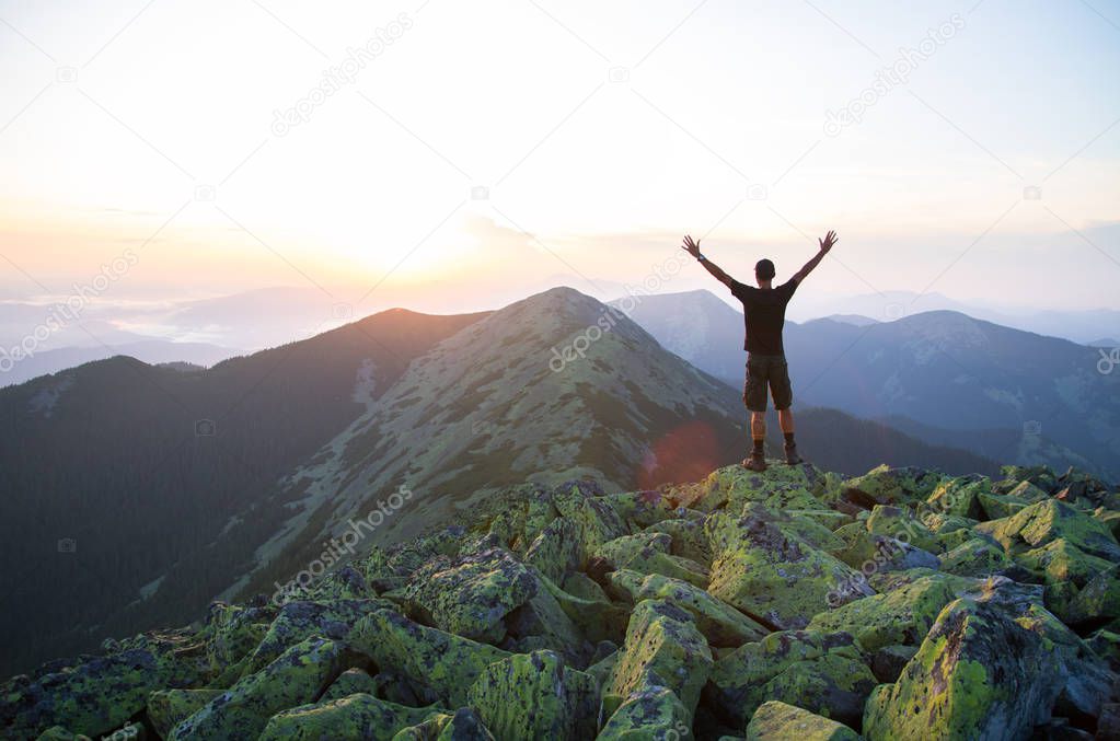caucasian man hiker is on the peak of green stones mountain watching sunset with hands in the air