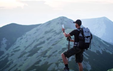 sırt çantası ve trekking çubukları ile kafkas adam yürüyüşçü yeşil taşların zirvesinde dağ bir akıllı telefon ile gün batımı fotoğrafları çekiyor
