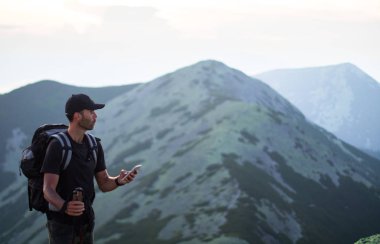 sırt çantası ve trekking çubukları ile kafkas adam yürüyüşçü yeşil taşların zirvesinde dağ akıllı telefon bir şey arıyor