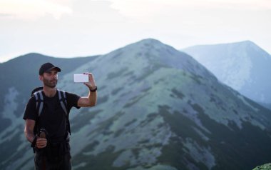 sırt çantası ve trekking çubukları ile kafkas adam yürüyüşçü yeşil taşların zirvesinde dağ bir akıllı telefon ile gün batımı fotoğrafları çekiyor
