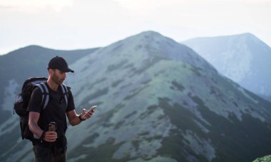 sırt çantası ve trekking çubukları ile kafkas adam yürüyüşçü yeşil taşların zirvesinde dağ akıllı telefon bir şey arıyor