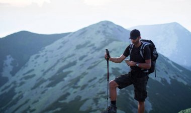 sırt çantası ve trekking çubukları ile kafkas adam yürüyüşçü yeşil taşların zirvesinde dağ akıllı telefon bir şey arıyor
