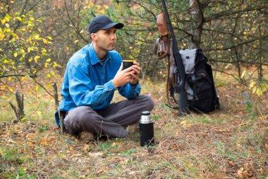 Beyaz erkek avcı ormanda dinleniyor. Termos çay içme. tüfek, cephane, arka planda sırt çantası