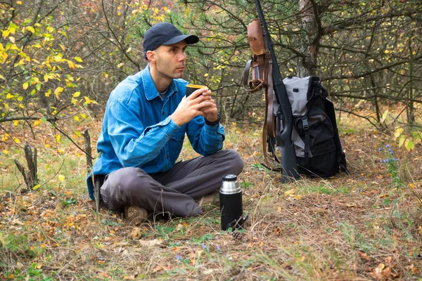白人男性ハンターが森で休んでいます。ポットからお茶を飲みます。散弾銃、弾薬、背景にバックパック — ストック写真